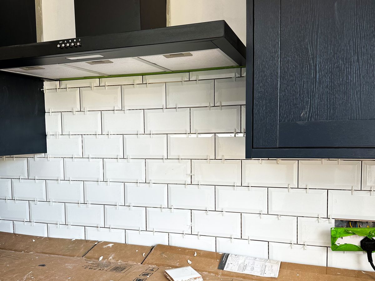 Metro wall tiles being fitted between navy kitchen units and a black cooker hood